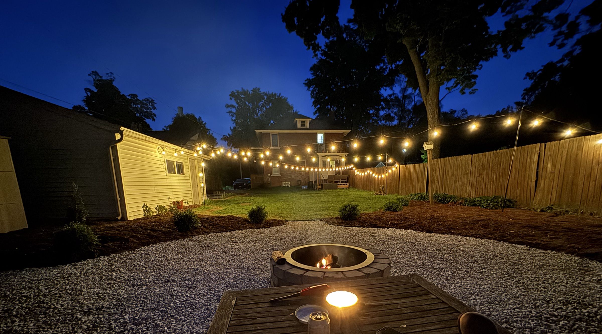 A backyard at dusk with string lights over a fire pit, freshly planted beds, and a lawn