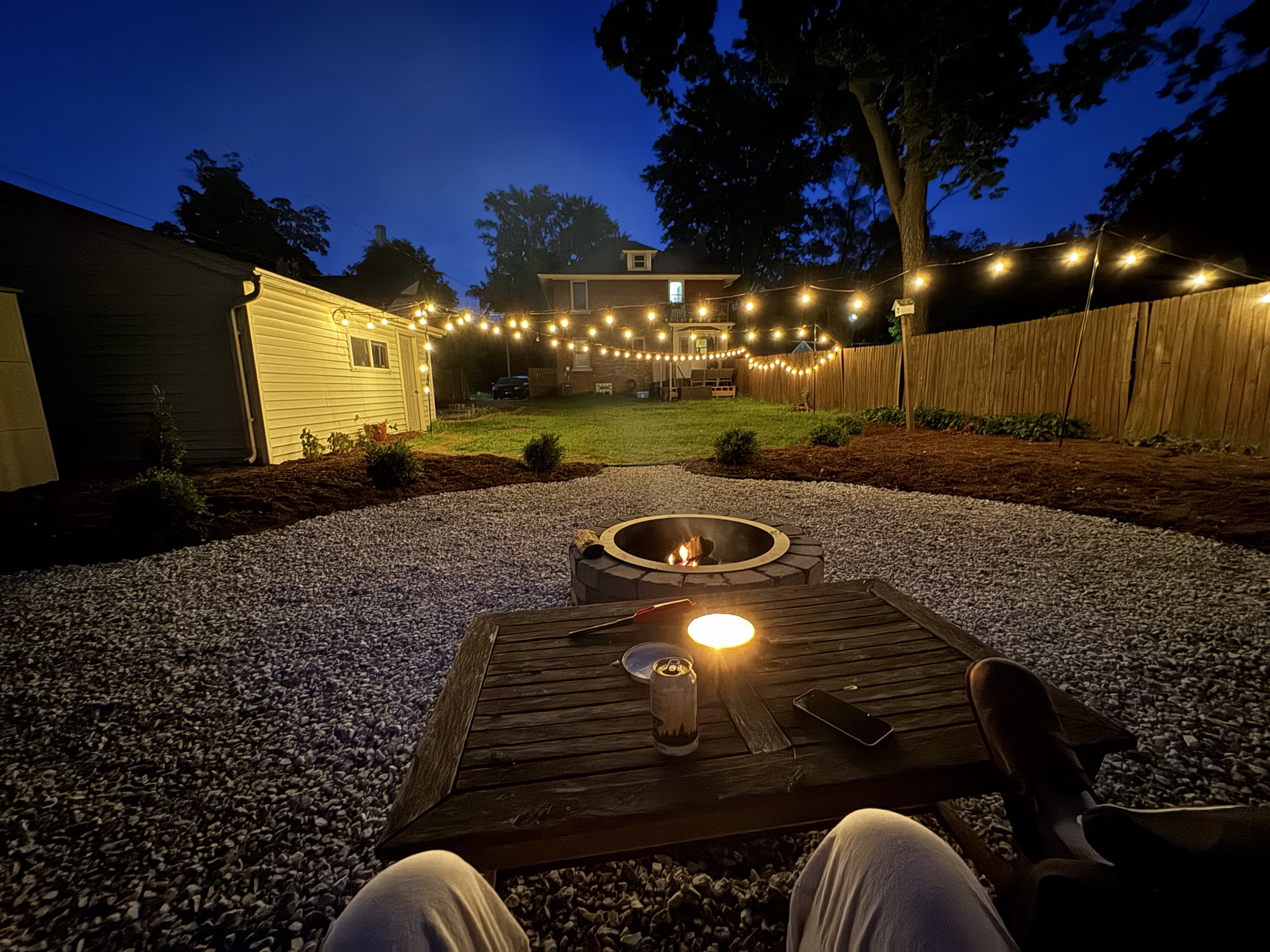 The same backyard at night, string lights glowing over the fire pit